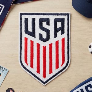 May include: A collection of USA-themed items, including a navy blue baseball cap, a scarf with red fringe, a patch, a soccer ball, and various pins and bracelets. The patch features the word "USA" in navy blue.