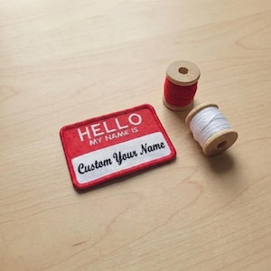 May include: A red and white embroidered name tag with the text "HELLO MY NAME IS" and a space to write "Custom Your Name". Two wooden spools of thread, one red and one white, are next to the name tag.