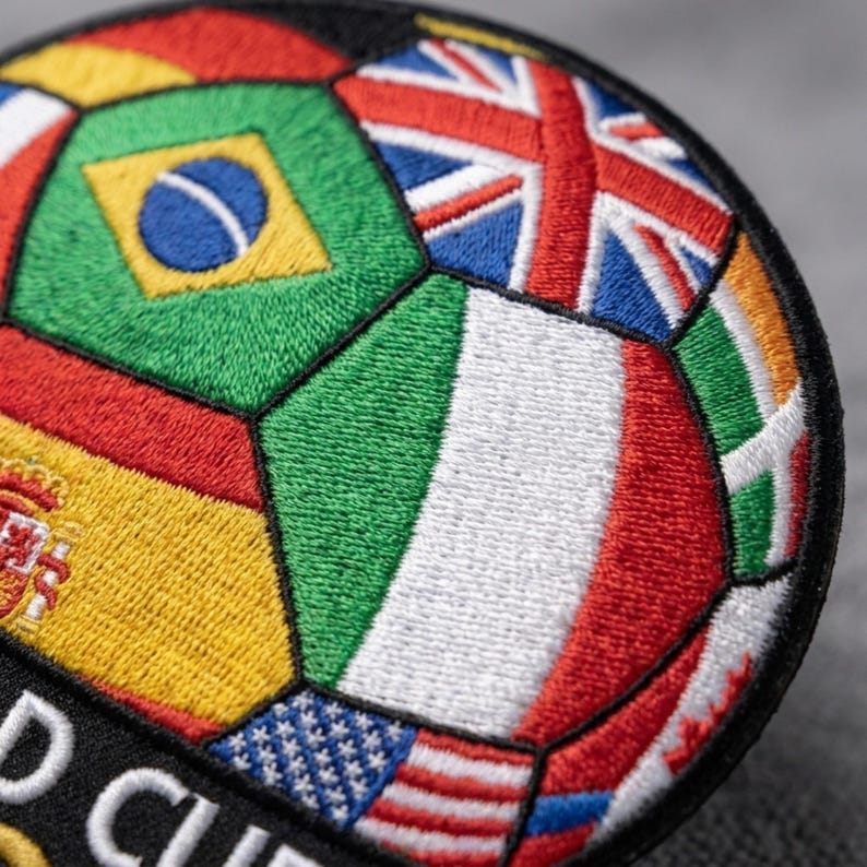 May include: Embroidered patch in the shape of a soccer ball, featuring the flags of various countries. The design includes the flags of the United States, Brazil, Italy, and the United Kingdom. The patch has a black border and the words "WORLD CUP" are visible.