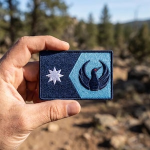 May include: A rectangular patch with a dark blue background and a light blue geometric design. The patch features a white star and a stylized bird with a red eye. The patch is held against a blurred outdoor background.