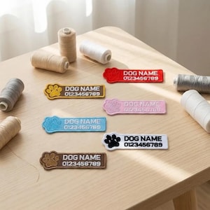 May include: Embroidered dog name tags in various colors, including red, pink, blue, yellow, brown, and white. Each tag features a paw print design and the text "DOG NAME" and a phone number. Spools of thread are in the background.