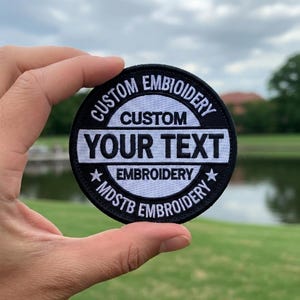 May include: A round, black embroidered patch with white text. The patch reads "CUSTOM EMBROIDERY" around the top and bottom edges. The center features "CUSTOM YOUR TEXT EMBROIDERY". Two stars flank the text "MDSTB EMBROIDERY".