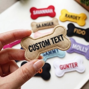 May include: A hand holding a beige bone-shaped patch with the text "CUSTOM TEXT" in black. Other bone-shaped patches in various colors, including red, yellow, black, and blue, are scattered around, each with a different name embroidered on them.