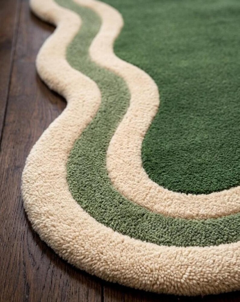 May include: A green, cream, and light green wavy rug. The rug has a soft, textured surface with a cream-colored border and two wavy stripes in light green and green. The rug is placed on a dark wood floor.