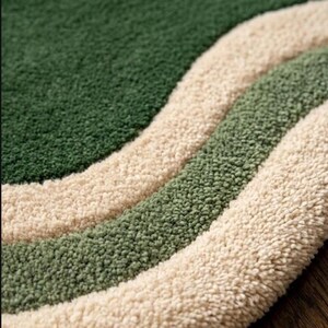 May include: Close-up of a textured rug with a wavy design. The rug features three distinct bands of color: a deep green, a lighter green, and a cream color. The rug has a soft, plush appearance.