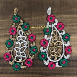May include: Pair of embroidered teardrop-shaped appliques. Each applique features a central design in gold and silver, surrounded by pink and green floral accents. The text "aum artisan" is visible.