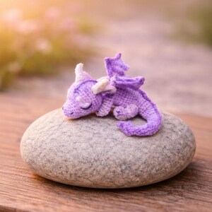 May include: A handcrafted, lavender-colored crochet dragon figurine with white accents, resting peacefully on a smooth, gray stone. The dragon has closed eyes and small wings. The background is blurred with soft, natural light.
