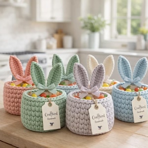 Puede incluir: Seis cestas de Pascua de ganchillo hechas a mano en colores pastel, cada una con orejas de conejo y un lazo. Las cestas están llenas de caramelos de colores y tienen etiquetas que dicen "Craftbird Handmade".