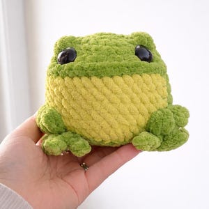 Peut inclure: Une peluche en forme de grenouille, faite à la main, dans des tons de vert et de jaune. La grenouille a de grands yeux noirs et est faite d'un fil doux et texturé. Un jouet mignon et câlin.