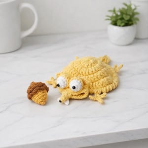 Puede incluir: Una criatura de ganchillo amarilla hecha a mano con grandes ojos blancos y una pequeña bellota marrón y amarilla. La criatura tiene patas pequeñas y un hocico largo. La bellota está al lado de la criatura sobre una superficie blanca.