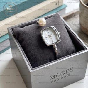 May include: A silver-toned quartz watch with a faceted square face and a white dial, paired with a single white pearl accent. The watch is displayed on a dark grey velvet cushion inside a square metallic grey box labelled MOSES RADIANCE.