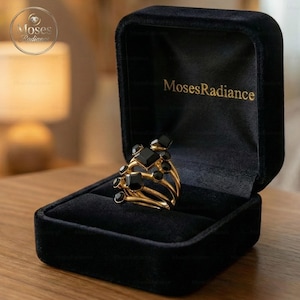 Könnte beinhalten: Ein goldfarbener Ring mit mehreren Bändern, verziert mit schwarzen geometrischen und runden Edelsteinen, präsentiert in einer offenen schwarzen Samt-Schmuckschatulle. Die Schatulle ist mit "Moses Radiance" geprägt.