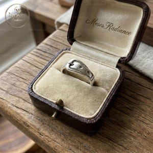 May include: A silver ring in the shape of a belt buckle, displayed in a vintage brown jewelry box with a beige interior. The ring has a polished finish and three small circular details. The box is open, revealing the ring.