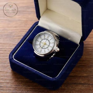May include: A silver ring watch with a white face and black numerals, presented in a blue velvet box. The watch has a quartz movement and a small crown. A unique accessory, the ring watch.