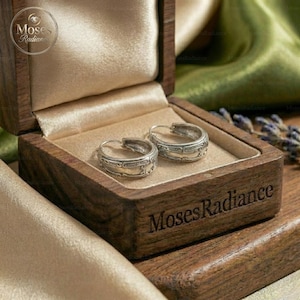 Puede incluir: Un par de pendientes de aro plateados con detalles intrincados se encuentra dentro de una caja de madera forrada con tela beige. La caja está grabada con las palabras "Moses Radiance". Los pendientes tienen un estilo vintage.