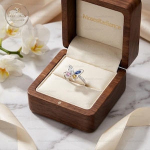 May include: A silver butterfly ring with pink, white, yellow, and blue gemstones, presented in an open wooden jewellery box. The box has a cream-coloured interior. The ring's design is the focal point within the box.