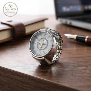 Puede incluir: Un reloj de anillo plateado con esfera blanca y manecillas doradas. El reloj tiene un movimiento de cuarzo y una banda el&aacute;stica. El reloj est&aacute; sobre una superficie de madera, con un bol&iacute;grafo y un port&aacute;til en el fondo.