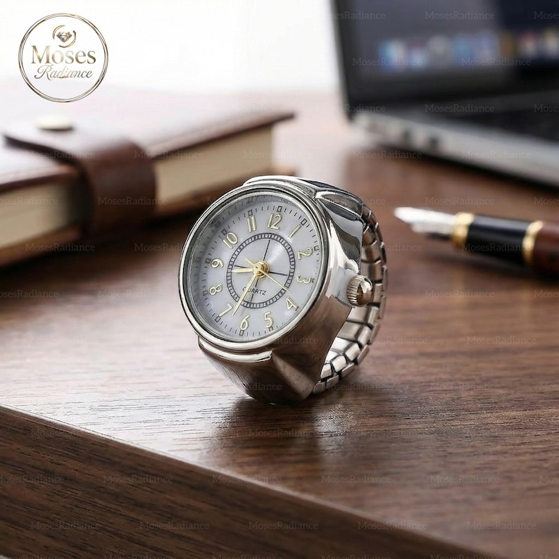 Puede incluir: Un reloj de anillo plateado con esfera blanca y manecillas doradas. El reloj tiene un movimiento de cuarzo y una banda el&aacute;stica. El reloj est&aacute; sobre una superficie de madera, con un bol&iacute;grafo y un port&aacute;til en el fondo.