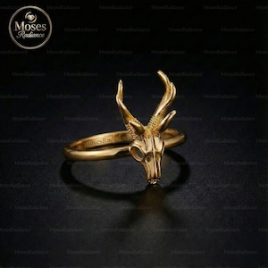 May include: A gold-colored ring featuring a deer skull design. The ring has a smooth band and a detailed skull with antlers. The ring is displayed on a dark surface, highlighting its intricate design. The ring is a unique piece of jewelry.