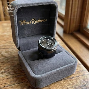 May include: A black ring watch with a dark grey face, presented in a grey velvet box. The watch features silver hands and hour markers. The box is embossed with "Moses Radiance" in gold lettering.