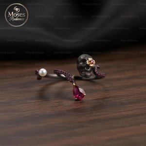 Puede incluir: Un anillo de plata oscura con un diseño de calavera, un acento floral y un rubí en forma de lágrima. El anillo también presenta una perla y pequeñas piedras preciosas rojas. El anillo está sobre un fondo oscuro.