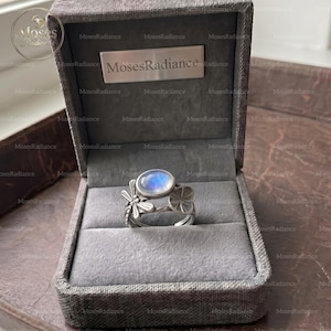 Peut inclure: Une bague en argent ornée d'un cabochon de pierre de lune ovale, agrémentée d'une libellule et d'un motif de feuille sur l'anneau. La bague est présentée dans un écrin à bijoux doublé de tissu gris avec une plaque en métal argenté MosesRadiance.