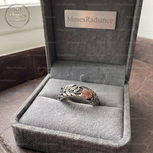 May include: A silver-toned ring featuring an embossed leaf and floral pattern with a copper-colored accent, presented inside a grey fabric-lined jewelry box with the text MosesRadiance printed on the interior lid and a circular logo.