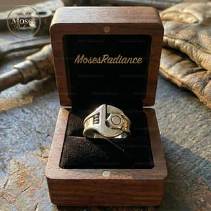 Puede incluir: Un anillo plateado con forma de llave inglesa y tornillo, presentado en una caja de madera abierta con interior de terciopelo negro. El nombre de la marca "Moses Radiance" es visible en la caja.