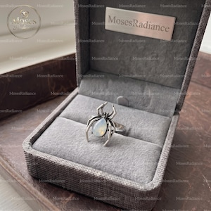 May include: A silver-toned ring shaped like a spider with a pear-shaped, iridescent white stone set in its abdomen. The ring is displayed inside a gray, textured, hinged jewelry box with a metal plaque reading MosesRadiance on the interior lid.