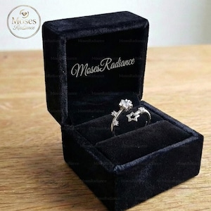 May include: A silver ring with a star and floral design, set in a black velvet ring box. The ring features a large, clear gemstone and is displayed open in the box. The box has the text "Moses Radiance" on the inside.