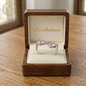 以下が含まれることがあります： 木製の箱に入った、ハート型のピンク色の宝石が2つ付いたシルバーリング。 箱は開いており、リングが見えています。 箱の内側にはブランド名「Moses Radiance」が刻まれています。