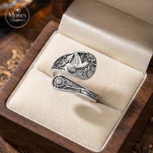 May include: A silver-colored ring with a hummingbird and sunflower design. The ring is adjustable and features detailed floral and bird motifs. It is presented in a wooden jewelry box with a soft, cream-colored interior.