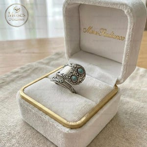 May include: A silver-toned ring with floral and scrollwork details, accented with small turquoise-colored stones. The ring is displayed in a white velvet ring box with gold trim and the text "Moses Radiance".