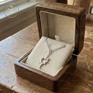 May include: A silver pendant necklace with a unique design, displayed inside an open wooden box with a soft, beige interior. The box is engraved with the text "MosesRadiance" on the lid. The necklace features a stylized heart shape.