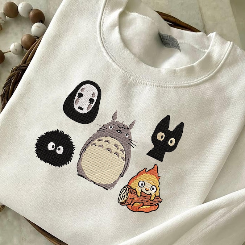 May include: White sweatshirt featuring embroidered cartoon characters, including a large gray Totoro, a black cat, a soot sprite, a No-Face spirit, and a fire demon. The design is centered on the front of the garment.
