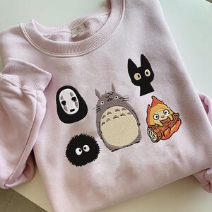 May include: A light pink sweatshirt featuring embroidered cartoon characters. The design includes a gray Totoro, a black cat, a soot sprite, a character from Spirited Away, and a fire demon. The sweatshirt has a crew neck and long sleeves.