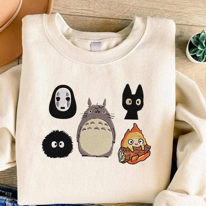 May include: Cream-colored sweatshirt featuring embroidered cartoon characters. The design includes a large, gray Totoro, a black cat, a soot sprite, a character with a white mask, and a fire-like creature.