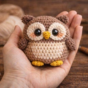 Puede incluir: Un búho de ganchillo hecho a mano en tonos marrón, crema y amarillo. El búho tiene grandes ojos negros, un pico amarillo y pequeñas plumas en las orejas. El búho se sostiene en una mano, mostrando su tamaño y detalle. Un artículo decorativo encantador y caprichoso.