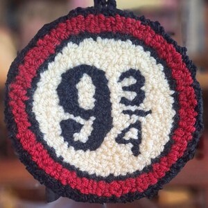 Puede incluir: Un tapiz circular de punch needle con un centro color crema y el texto negro 9 3/4. El diseño está bordeado por un anillo rojo grueso y un borde exterior negro, creando una apariencia texturizada y artesanal con bucles de hilo visibles.
