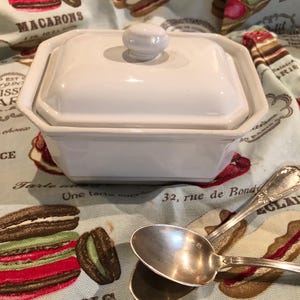 Può includere: Un piatto in ceramica bianco, ottagonale, con coperchio abbinato e piccola maniglia rotonda. Due cucchiai d'argento antichi sono accanto al piatto. Il piatto è su un tessuto azzurro con immagini di pasticceria e testo francese.