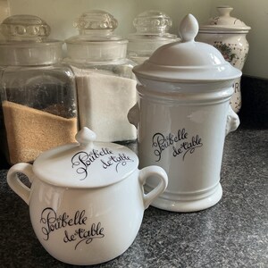 Könnte beinhalten: Weiße Keramikbehälter mit Deckeln, mit dem Schriftzug "Poubelle de table" in Schwarz. Ein Behälter ist rund mit Griffen, der andere zylindrisch. Im Hintergrund befinden sich mehrere Gläser mit Deckeln.