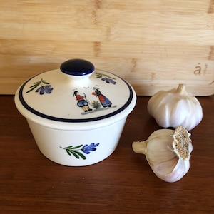 Puede incluir: Un tarro de cerámica blanco para guardar ajos con borde y tapa azules. La tapa tiene un pomo azul y una escena pintada de dos figuras. Dos cabezas de ajo están junto al tarro sobre una superficie de madera.