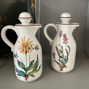 May include: Two white ceramic pitchers with floral designs. One pitcher features a yellow and orange flower with green leaves, the other has purple flowers. Both have a handle, spout, and a lid with a brown trim.