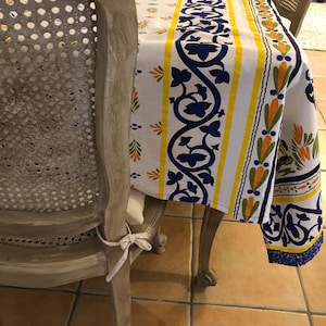 Op de afbeelding: Een wit tafelkleed met een blauw bloemmotief en gele strepen. Het tafelkleed ligt op een houten tafel. Het ontwerp omvat blauwe bloempatronen en oranje accenten. Er staat een stoel met rieten rug naast.