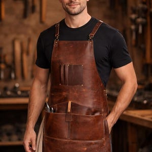 May include: A brown leather apron with multiple pockets and adjustable straps. The apron is worn over a black t-shirt. The apron has a rugged, worn appearance, suggesting durability and craftsmanship. The apron is designed for work or craft activities.