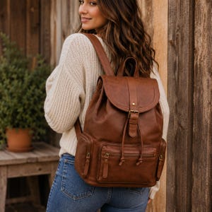 May include: A medium brown backpack featuring a fold-over flap with a buckle closure, a drawstring main compartment, and three exterior zippered pockets. The bag includes adjustable shoulder straps and a top handle, crafted with a smooth finish.