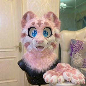 Op de afbeelding: Een roze en witte harige fursuit-kop en bijpassende pootkussens. De kop heeft grote blauwe ogen met hartvormige pupillen, een roze neus en een lachende uitdrukking met de tong uit. De pootkussens zijn wit met roze kussens.