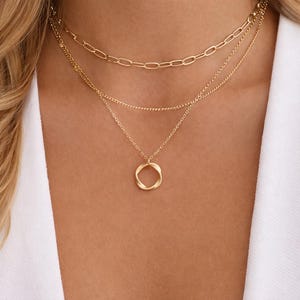 May include: A set of three gold necklaces. The top necklace has a chain link design, the middle one is a fine chain, and the bottom one features a twisted circle pendant. The necklaces are shown against a white background.