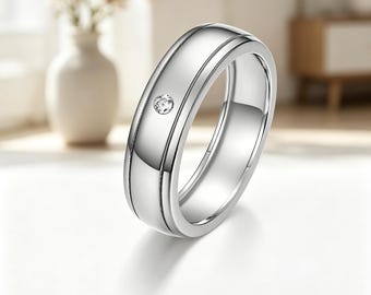 Puede incluir: Un anillo de banda en tono plateado pulido con una superficie lisa y redondeada, que presenta una pequeña piedra preciosa transparente de corte redondo engastada en el centro. Tiene dos ranuras paralelas en la circunferencia.