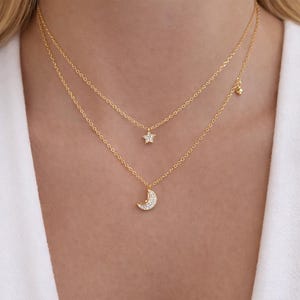 Può includere: Due delicate collane d'oro. La collana superiore presenta un piccolo ciondolo a forma di stella, e la collana inferiore un ciondolo a forma di mezzaluna. Entrambi i ciondoli sono incastonati con piccole pietre scintillanti. Le collane sono sovrapposte.
